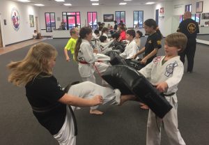 Kids training on taekwondo kicks with safety shields during a KFMA family martial arts class.